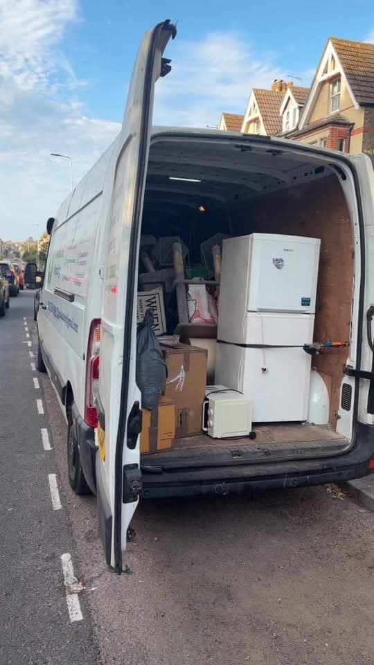 The back of a van open on a street, loaded with a fridge freezer, boxes and furniture
